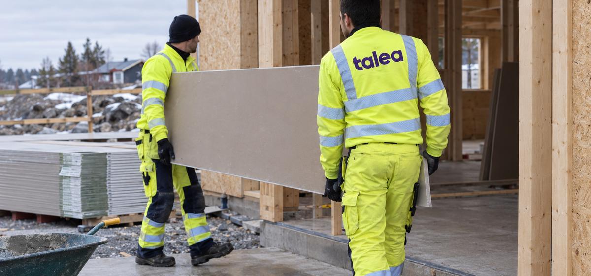 Två Talea-medarbetare bär in en gipsskiva på ett bygge